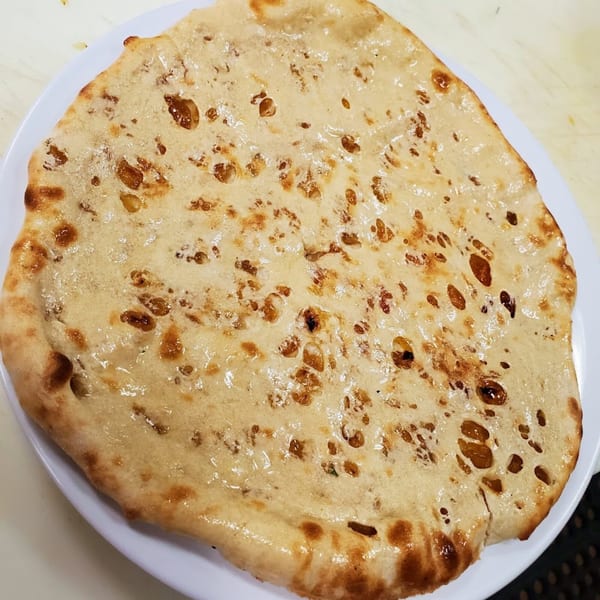 Close-up of a large, round, golden-brown naan bread with bubbly texture on a white plate.