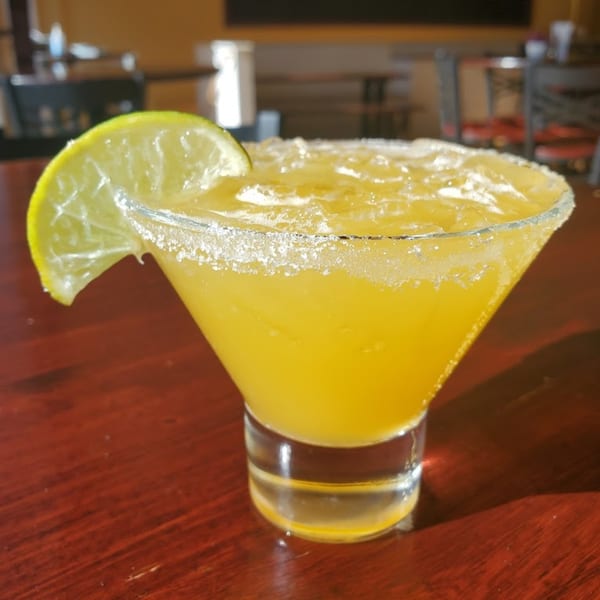 A bright yellow margarita with a salted rim and lime wedge, on a dark wooden table.