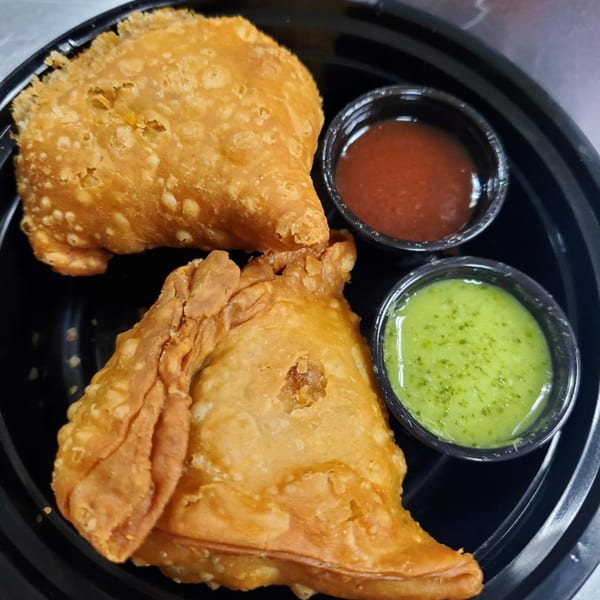 Two golden-brown samosas with small cups of red and green chutneys in a black container.